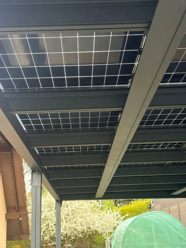 Vue en contre-plongée sous une pergola solaire, jardin arboré et petite serre en arrière-plan.