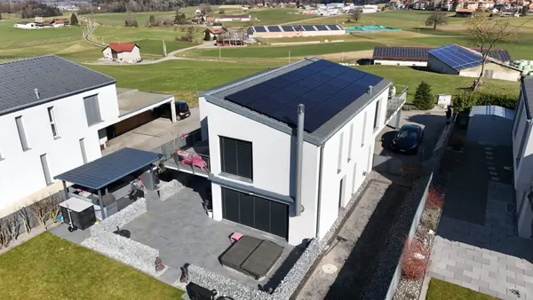 Maison moderne à toit plat avec panneaux solaires, campagne vallonnée et fermes au loin.