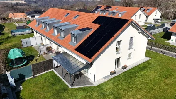 Long bâtiment résidentiel à toiture orangée avec panneaux noirs, jardin et trampoline attenants.