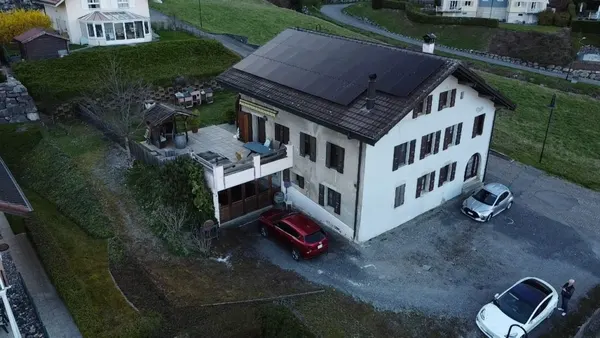 Grande maison ancienne aux volets foncés, panneaux solaires en toiture, coteau en arrière-plan.