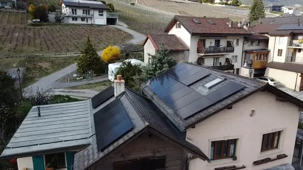 Toiture couverte de panneaux noirs sur un hameau de montagne, vignes en terrasses visibles.