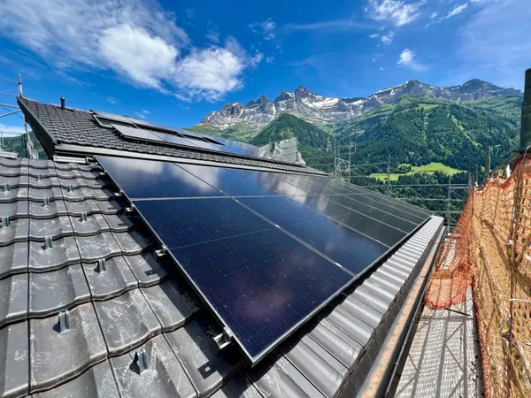 Panneaux solaires sur toiture à tuiles, sommets rocheux escarpés et vallée verdoyante en arrière-plan.